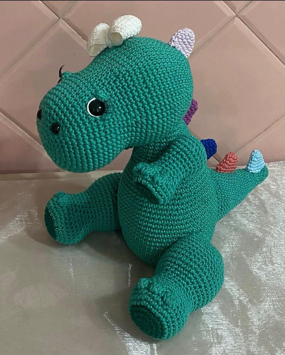 Amigurumi de dinossauro verde com espinhos coloridos nas costas e laço branco na cabeça.