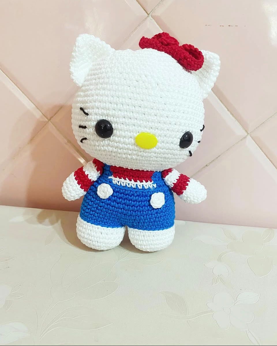 Amigurumi de gatinha branca com laço vermelho e macacão azul em crochê.
