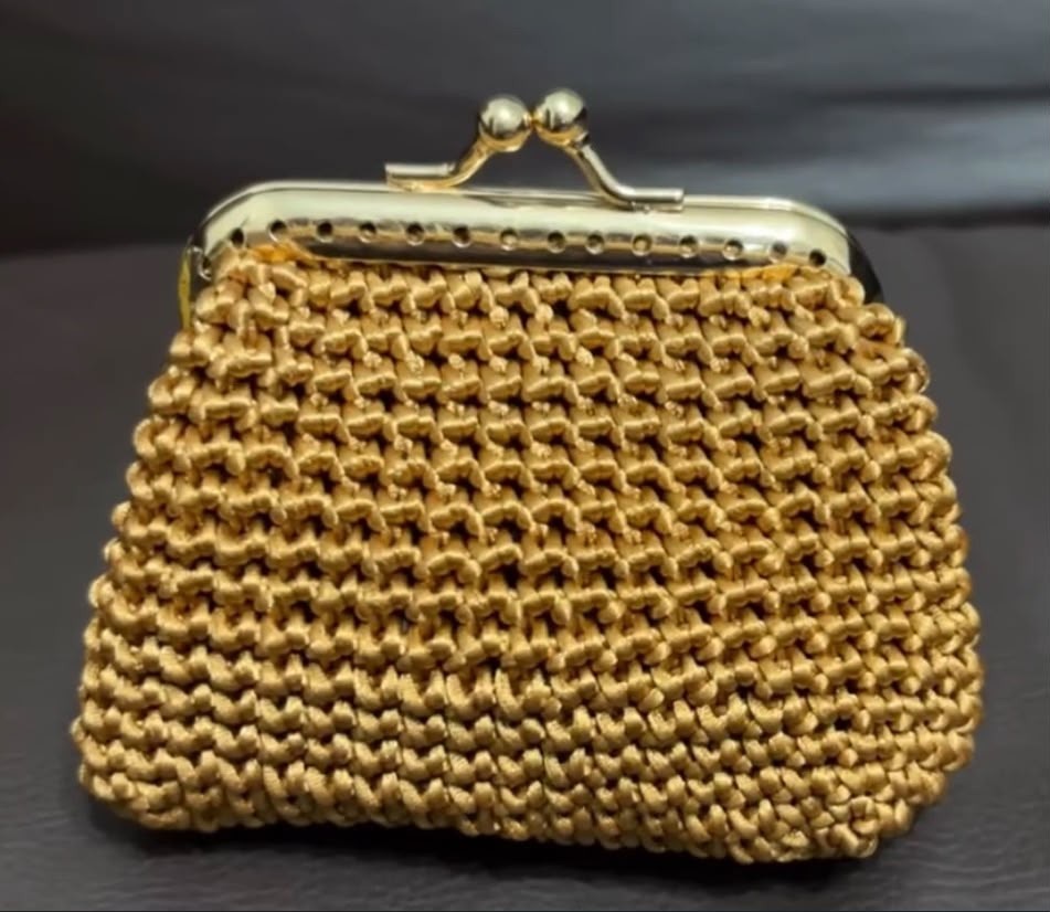 Porta-níquel dourado em crochê com fecho da vovó de duas esferas metálicas e alça de corrente, apoiado sobre superfície escura.