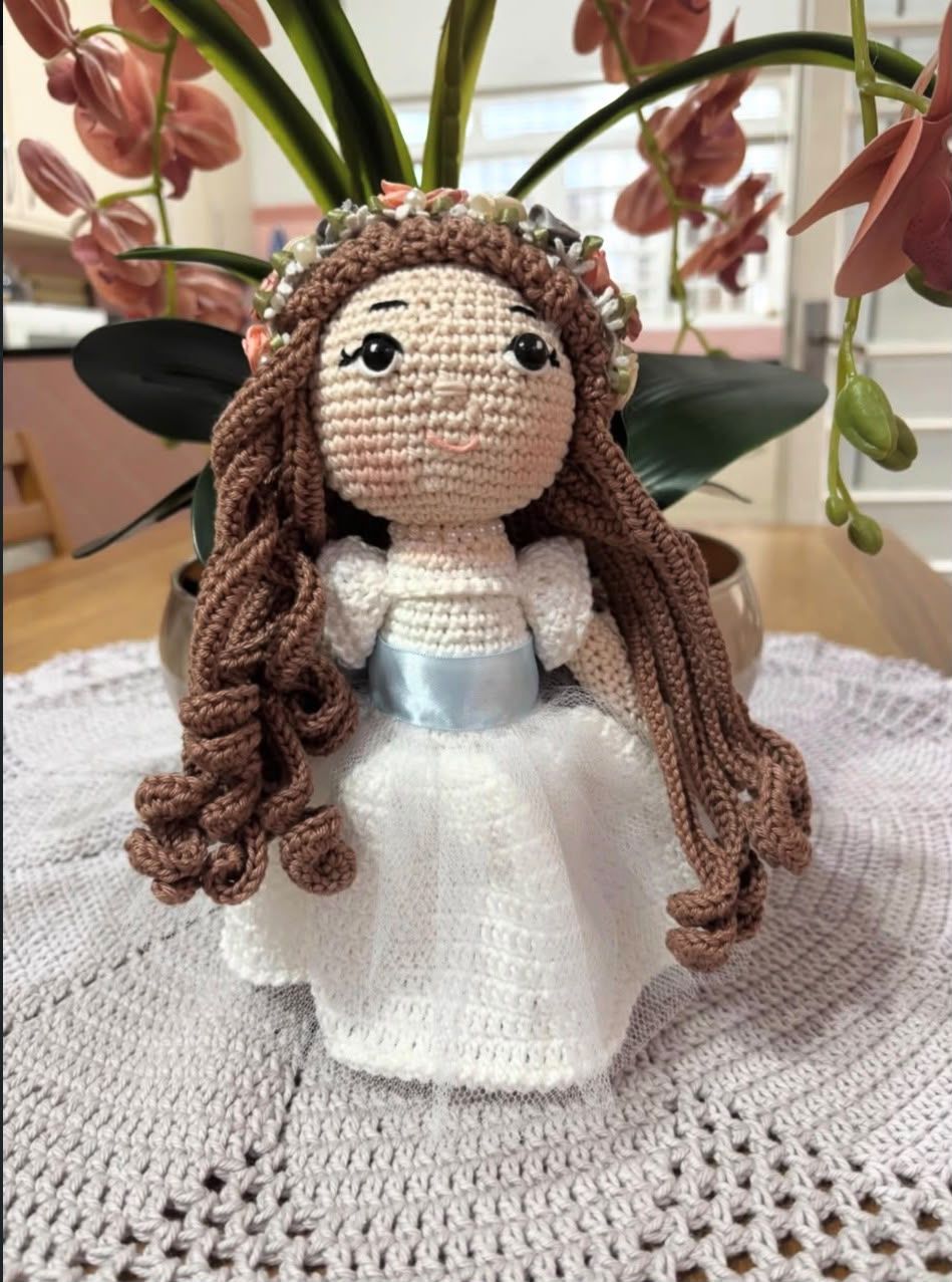 Boneca de noiva em crochê, de vestido branco com saia de tule e coroa de flores, apoiada sobre toalha de crochê cinza.