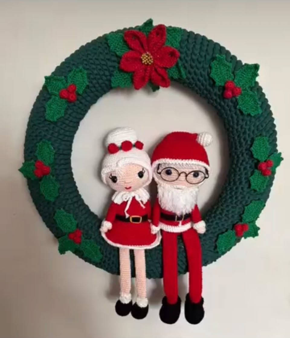 Guirlanda de Natal verde em crochê com flor de Natal vermelha, folhas de azevinho e o casal Papai e Mamãe Noel pendurado no centro.