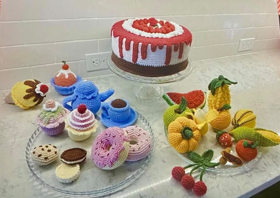 Bolo de aniversário, cupcakes, donuts, chá e frutas gigantes confeccionados em crochê sobre bancada de cozinha.