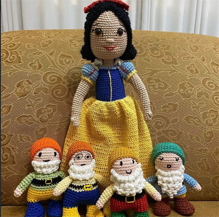 Personagem de conto de fadas em crochê com vestido azul e amarelo, acompanhada de quatro bonecos de anões com barba branca e gorros coloridos.