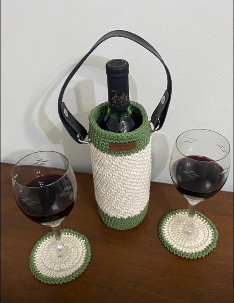 Porta-garrafa de vinho em crochê verde e off-white, com alça escura de couro, vestindo uma garrafa, ao lado de duas taças de vinho tinto e dois descansos de copo combinando.