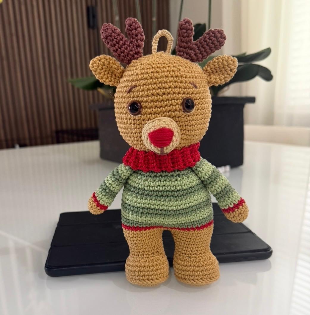 Rena de Natal em crochê, em fio bege, com chifres marrons, blusa verde de punhos vermelhos, gola alta vermelha e alça no alto da cabeça, apoiada em pé sobre mesa clara.