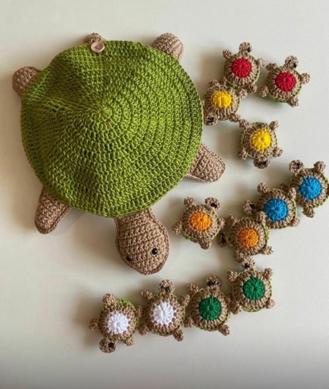 Tartaruga grande em crochê com casco verde e corpo bege, com botão no alto do casco, cercada por mini tartarugas de cascos vermelho, amarelo, laranja, azul, verde e branco.